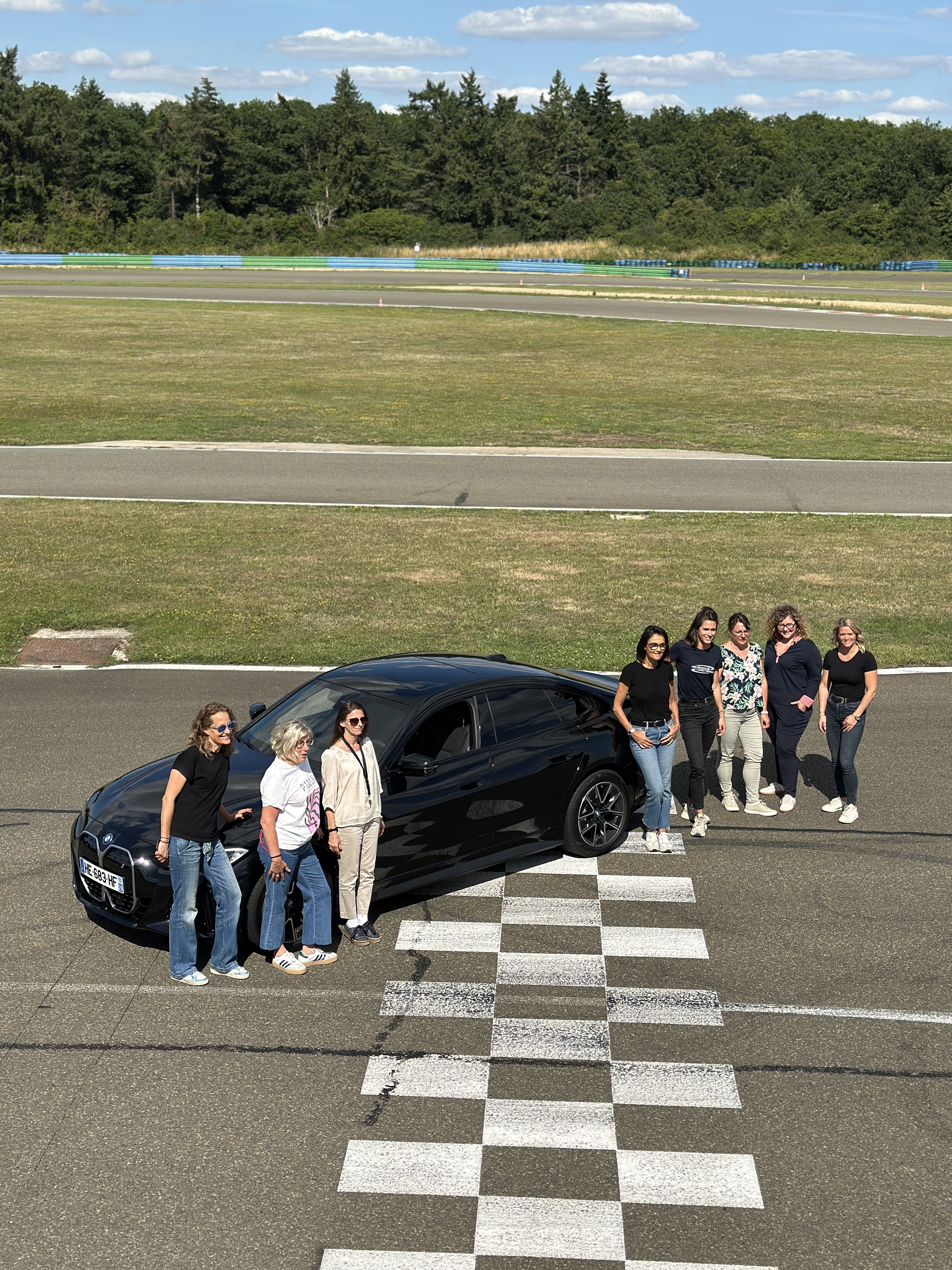 Groupe d'une dizaine de participantes en demi-cercle devant une BMW i4 sur le damier du circuit, lors d'une journée 100 % féminine encadrée par Nina.